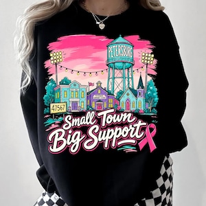 May include: A black sweatshirt featuring a colorful graphic of a small town scene with a water tower labeled Petersburg, a city hall, a school, and a church. The text reads Small Town Big Support with a pink awareness ribbon on the right side.