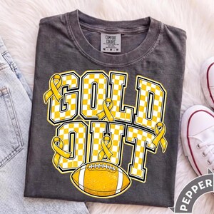 Puede incluir: Una camiseta gris carb&oacute;n de manga corta con el texto GOLD OUT en un patr&oacute;n de cuadros amarillos y blancos. El dise&ntilde;o incluye cintas de concienciaci&oacute;n amarillas integradas en las letras y un gr&aacute;fico de bal&oacute;n de f&uacute;tbol americano en el centro.