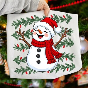 Puede incluir: Una camiseta blanca con una ilustraci&oacute;n gr&aacute;fica de un mu&ntilde;eco de nieve sonriente que lleva un gorro de Pap&aacute; Noel rojo y una bufanda de punto roja. El mu&ntilde;eco tiene brazos de rama y tres botones negros sobre un fondo de ramas de pino.