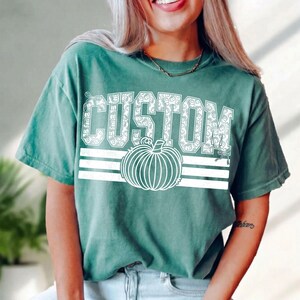 May include: A sage green short-sleeve t-shirt featuring the word CUSTOM in white block letters filled with a small pumpkin pattern. Below the text is a large white line-art pumpkin graphic centered over three horizontal white stripes.