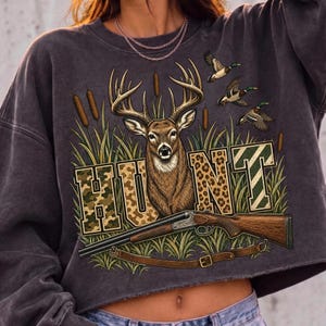 May include: A charcoal grey crewneck sweatshirt featuring a graphic print of a deer with antlers, three flying ducks, and bulrushes. The word HUNT is printed in large block letters with camouflage and leopard patterns. A shotgun is positioned below.