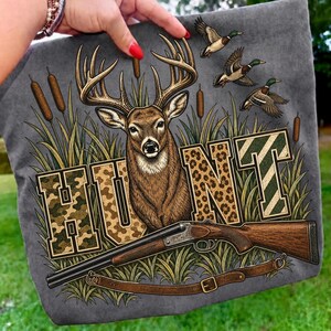 May include: A charcoal grey t-shirt featuring a graphic print of a deer with large antlers, three flying ducks, and a shotgun. The word HUNT is displayed in large block letters with camouflage and leopard print patterns amidst tall grass.