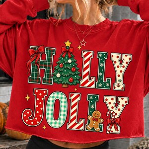 Peut inclure: Un sweat-shirt rouge &agrave; manches longues avec le texte HOLLY JOLLY en grandes lettres d&eacute;coratives. Les lettres sont remplies de motifs incluant du tartan vert, des rayures rouges et blanches et des pois rouges. Un sapin vert et un bonhomme en pain d'&eacute;pice.