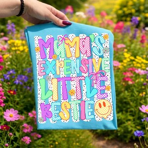 May include: A bright blue t-shirt featuring a white background with black vertical stripes and dots. The text reads MAMA'S EXPENSIVE LITTLE BESTIE in colorful, patterned bubble letters with small pink bows and a yellow smiley face icon.