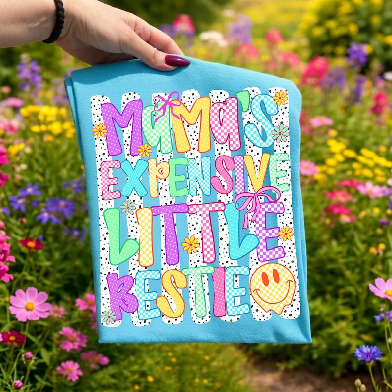 May include: A bright blue t-shirt featuring a white background with black vertical stripes and dots. The text reads MAMA'S EXPENSIVE LITTLE BESTIE in colorful, patterned bubble letters with small pink bows and a yellow smiley face icon.