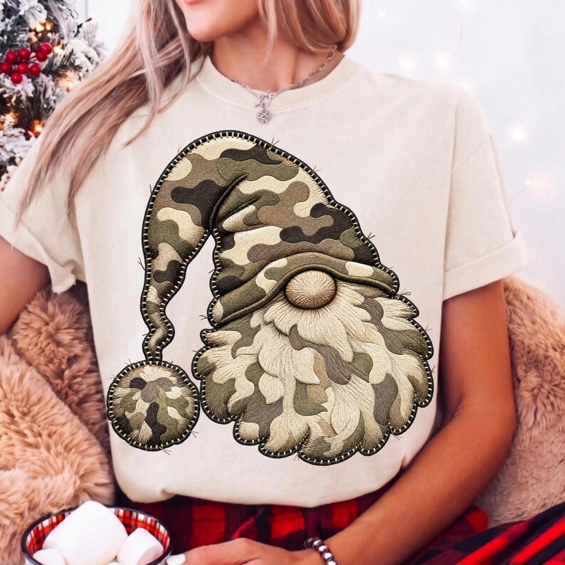 K&ouml;nnte beinhalten: Ein cremefarbenes Kurzarm-T-Shirt mit einer gro&szlig;en Grafik eines Gnoms, der eine M&uuml;tze mit Tarnmuster tr&auml;gt. Der Gnom hat einen strukturierten, hellen Bart und einen passenden Tarn-Bommel am Ende seiner langen, herabh&auml;ngenden M&uuml;tze.