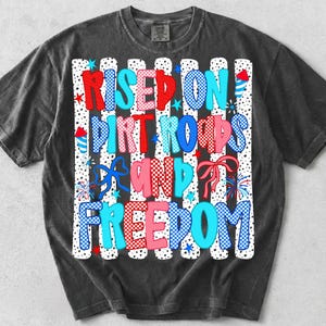 May include: A charcoal grey short-sleeve t-shirt featuring a graphic print with the text "RISED ON DIRT ROADS AND FREEDOM" in red, blue, and pink checkered and polka-dot patterns, accented with illustrations of fireworks, bows, and stars.