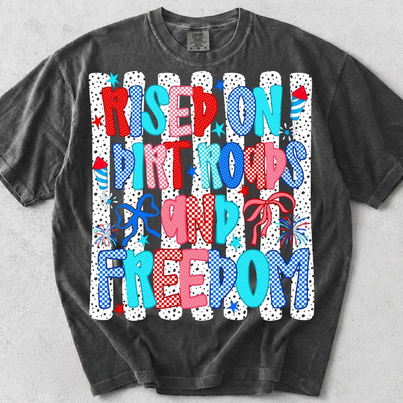 May include: A charcoal grey short-sleeve t-shirt featuring a graphic print with the text "RISED ON DIRT ROADS AND FREEDOM" in red, blue, and pink checkered and polka-dot patterns, accented with illustrations of fireworks, bows, and stars.