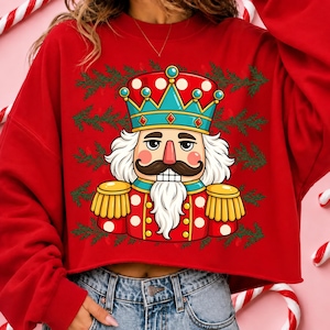 Puede incluir: Una sudadera roja de manga larga con un estampado gr&aacute;fico de un cascanueces con corona, bigote y barba blanca. El dise&ntilde;o est&aacute; rodeado por patrones de ramas de pino verdes. La sudadera tiene un dobladillo corto y cuello redondo.