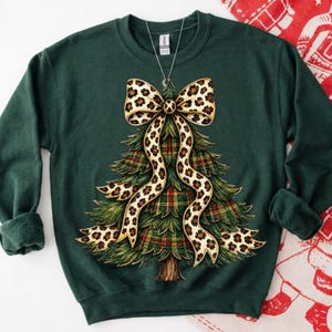 May include: A dark green long-sleeve sweatshirt featuring a graphic of a Christmas tree with a plaid pattern and a large leopard print bow draped over the top. The design includes a brown trunk at the base and a silver chain necklace hanging above.