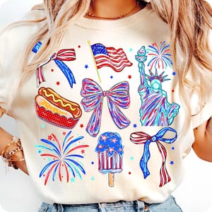 May include: A cream-colored t-shirt featuring a patriotic graphic print with a hot dog, the Statue of Liberty, fireworks, an American flag, and three decorative bows in red, white, and blue colors, accented with small star shapes throughout.