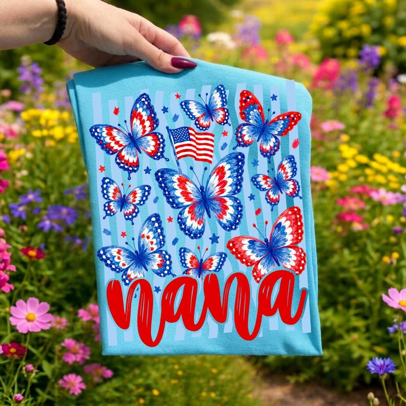 May include: A light blue t-shirt featuring a graphic print of multiple butterflies with red, white, and blue wing patterns. A small American flag is positioned near the center butterfly. The word "nana" is printed in large red script at the bottom.
