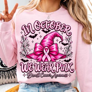 May include: A light pink sweatshirt featuring a graphic of a pink witch hat adorned with a pink ribbon. The text reads "IN OCTOBER WE WEAR PINK" and "Breast Cancer Awareness" with decorative black bats and floral flourishes surrounding the central design.
