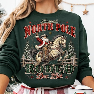May include: A dark green crewneck sweatshirt featuring a graphic of Santa Claus riding a horse. The text reads Annual NORTH POLE RODEO Dec. 25th. The design includes pine trees, a wooden fence, and a lasso. The sweatshirt has long sleeves.