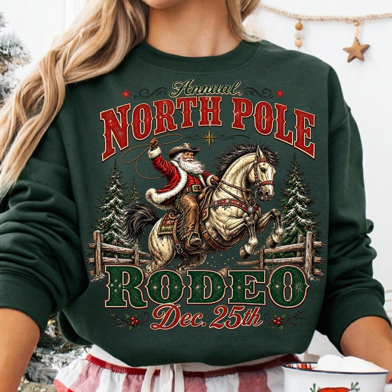 May include: A dark green crewneck sweatshirt featuring a graphic of Santa Claus riding a horse. The text reads Annual NORTH POLE RODEO Dec. 25th. The design includes pine trees, a wooden fence, and a lasso. The sweatshirt has long sleeves.