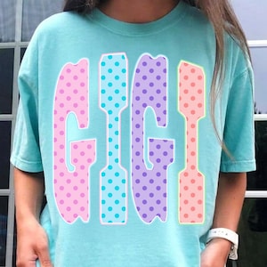 May include: A light blue t-shirt with the word "GIGI" in large, colorful letters. The letters are outlined in white and filled with polka dots in pink, blue, purple, and peach. The shirt has short sleeves and a crew neck.