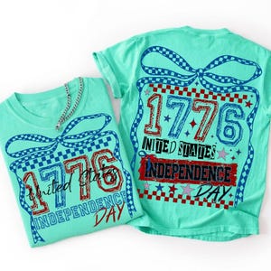 Może przedstawiać: Dwie miętowe koszulki z grafiką niebieskiej kokardy w kratkę oraz napisem 1776 United States Independence Day w kolorze czerwonym i niebieskim. Wz&oacute;r zawiera gwiazdki i obw&oacute;dkę w kratkę, a do jednej koszulki przymocowany jest srebrny łańcuszek.