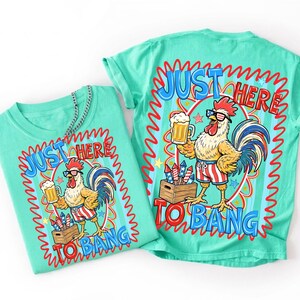 Op de afbeelding: Een mintgroen t-shirt met een afbeelding van een haan in een korte broek met Amerikaanse vlagpatronen en een zonnebril, die een bierpul vasthoudt naast een kist met vuurwerk. De tekst "JUST HERE TO BANG" staat in blauw en rood rond de illustratie.
