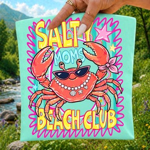 May include: A light mint green fabric pouch featuring a graphic of a red crab wearing black sunglasses and a pearl necklace with a seashell charm. The text reads SALTY MOMS BEACH CLUB in yellow letters, surrounded by pink squiggly lines and stars.