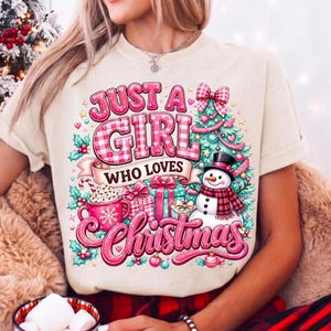 May include: A cream-colored t-shirt featuring a festive graphic with the text "JUST A GIRL WHO LOVES Christmas". The design includes a decorated Christmas tree, a snowman with a plaid scarf, wrapped gifts, and a pink mug with a snowflake.
