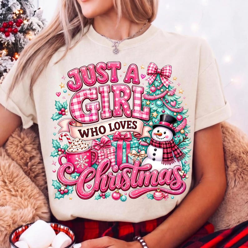 May include: A cream-colored t-shirt featuring a festive graphic with the text "JUST A GIRL WHO LOVES Christmas". The design includes a decorated Christmas tree, a snowman with a plaid scarf, wrapped gifts, and a pink mug with a snowflake.