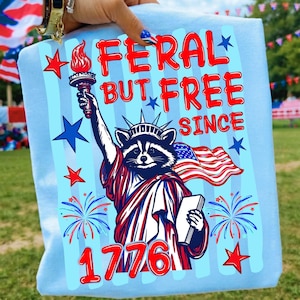 Może przedstawiać: Jasnoniebieski t-shirt z grafiką szopa pracza przebranego za Statuę Wolności, trzymającego pochodnię i książkę. Napis głosi FERAL BUT FREE SINCE 1776. Wz&oacute;r zawiera czerwone i niebieskie gwiazdy oraz motywy sztucznych ogni.