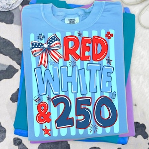 Peut inclure: Un t-shirt bleu clair avec une impression graphique portant le texte RED WHITE & 250 en rouge, bleu clair et bleu fonc&eacute;. Le motif comprend un n&oelig;ud patriotique, des &eacute;toiles et des feux d'artifice sur des rayures verticales bleu clair.