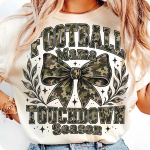 May include: A cream-coloured t-shirt featuring a large graphic with the text FOOTBALL MAMA TOUCHDOWN SEASON in a camouflage pattern. The design includes a central camouflage bow and leafy branch accents, all rendered in shades of green and brown.