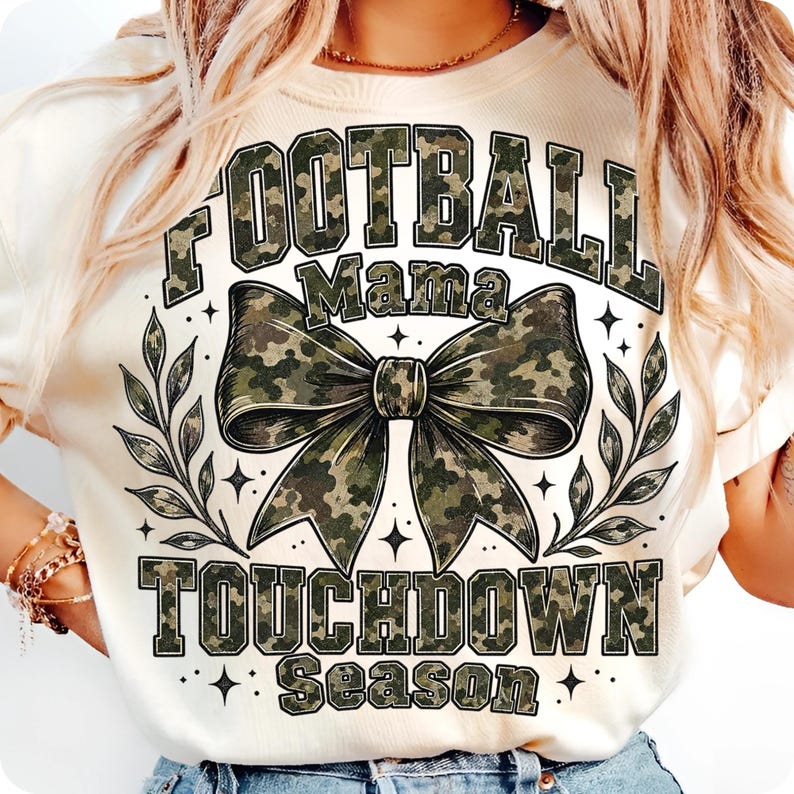 May include: A cream-coloured t-shirt featuring a large graphic with the text FOOTBALL MAMA TOUCHDOWN SEASON in a camouflage pattern. The design includes a central camouflage bow and leafy branch accents, all rendered in shades of green and brown.