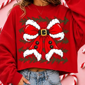 Puede incluir: Una sudadera roja de manga larga con un gran gr&aacute;fico de un lazo rojo adornado con piel sint&eacute;tica blanca. El lazo tiene un cintur&oacute;n negro con una hebilla dorada en el centro y cuatro botones negros en los extremos. Ramas de pino verdes rodean el lazo.