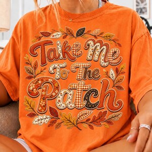 May include: An orange t-shirt featuring a graphic print with the text "Take Me To The Patch" in a bold, rounded font. The letters are filled with various patterns including gingham, floral, and solid colors, surrounded by a wreath of autumn leaves.