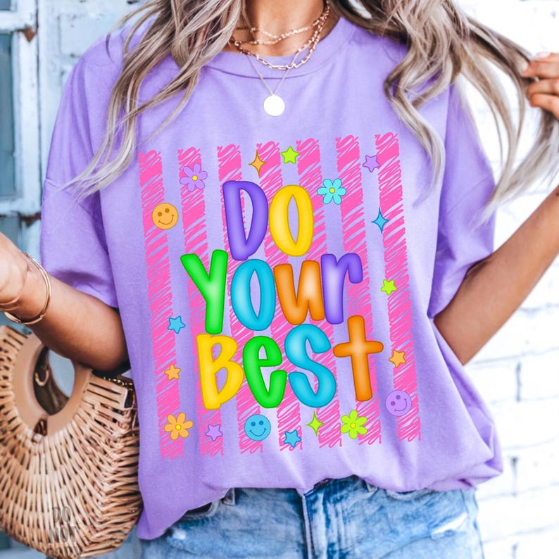 May include: A lilac-colored t-shirt with the words "DO YOUR BEST" in colorful, bubble letters. The text is set against a pink striped background, adorned with flowers, stars, and smiley faces. The shirt is a casual, everyday wear item.