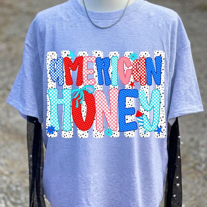 May include: A light gray short-sleeve t-shirt featuring a graphic print with the text AMERICAN HONEY. The letters are filled with red, blue, and pink patterns including polka dots and checkers, set against a background of vertical black dots.