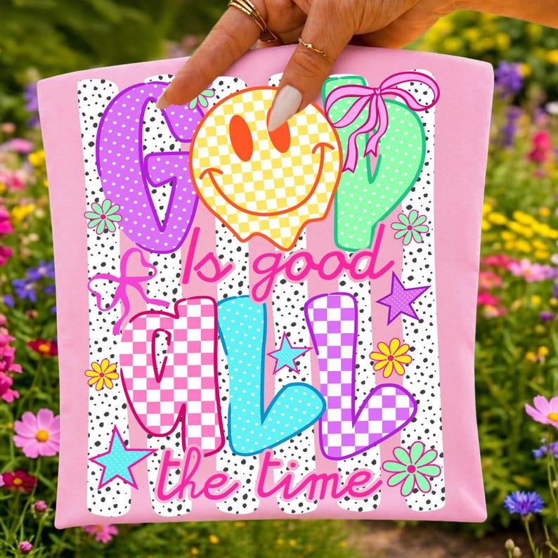 May include: A light pink fabric item featuring a graphic print with the text God Is Good All The Time. The design includes a yellow checkered smiley face, colorful checkered letters, pink bows, small stars, and flowers over a black dotted stripe pattern.