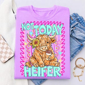 May include: A lilac-colored t-shirt featuring a cartoon Highland cow with a sunflower and coffee cup. The text "NOT TODAY HEIFER" is in turquoise and white. The design includes pink and yellow stars and a pink border.