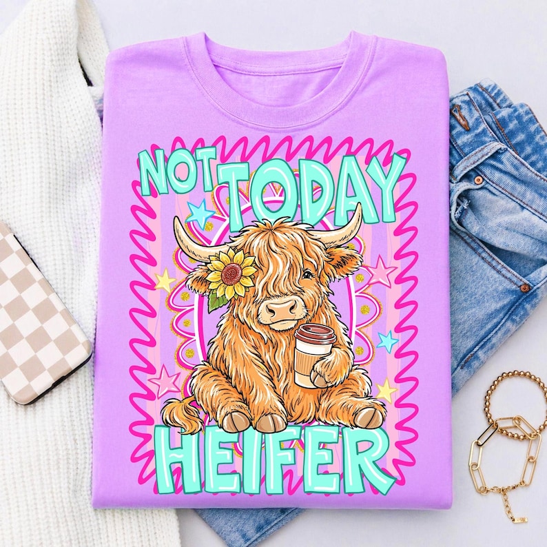 May include: A lilac-colored t-shirt featuring a cartoon Highland cow with a sunflower and coffee cup. The text "NOT TODAY HEIFER" is in turquoise and white. The design includes pink and yellow stars and a pink border.