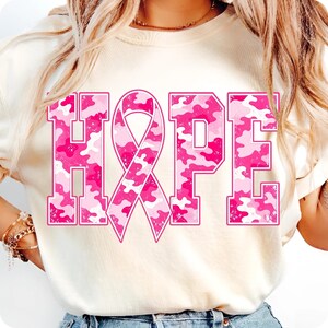 Peut inclure: Un t-shirt couleur cr&egrave;me avec le mot HOPE en grandes lettres majuscules. La lettre O est remplac&eacute;e par un ruban de sensibilisation rose. Le texte et le ruban pr&eacute;sentent un motif camouflage rose et blanc avec une texture vieillie.