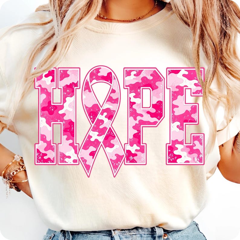 Peut inclure: Un t-shirt couleur cr&egrave;me avec le mot HOPE en grandes lettres majuscules. La lettre O est remplac&eacute;e par un ruban de sensibilisation rose. Le texte et le ruban pr&eacute;sentent un motif camouflage rose et blanc avec une texture vieillie.