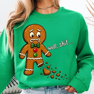 May include: A bright green crewneck sweatshirt featuring a graphic of a brown gingerbread person with a worried expression, a green bow tie, and red and green buttons. The gingerbread person has a missing foot with crumbs, and the text well, shit...