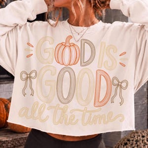 May include: A cream-colored long-sleeve sweatshirt featuring the text "GOD IS GOOD all the time" in a multi-line graphic. The design includes a central orange pumpkin illustration replacing the letter O in GOD, and two brown line-art bows on the sides.