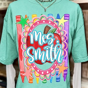 May include: A mint green short-sleeve t-shirt featuring a graphic print of a red apple inside a pink scalloped frame, surrounded by colorful crayon illustrations and stars. The text "Mrs. Smith" is printed in white script with a blue outline.
