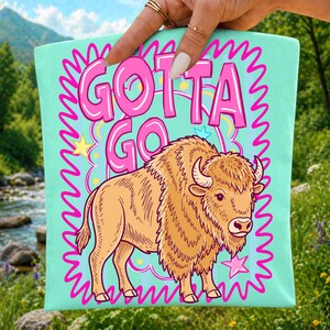 May include: A light mint green tote bag featuring a graphic illustration of a brown bison. Above the bison, the text "GOTTA GO" is printed in bold pink letters. The design is framed by a jagged pink border and includes small yellow and pink stars.