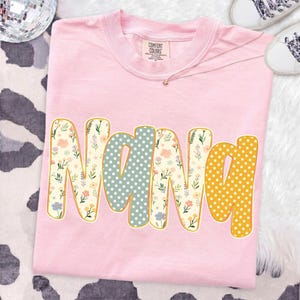 May include: A light pink t-shirt with the word NANA printed in large, rounded letters. The letters feature a mix of floral patterns in cream, blue, and yellow, alongside a star pattern in sage green and yellow, all outlined in a thin gold border.