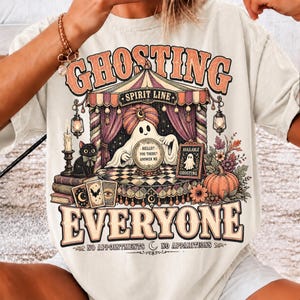 Pode incluir: Uma t-shirt cor de creme com um gr&aacute;fico de um fantasma de turbante atr&aacute;s de uma bola de cristal, com o texto "GHOSTING EVERYONE" e "HELLO? YOU THERE? ANSWER ME". O design inclui um gato preto, cartas de tarot e uma ab&oacute;bora.