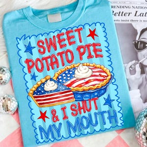 Op de afbeelding: Een lichtblauw t-shirt met een afbeelding met de tekst "SWEET POTATO PIE & I SHUT MY MOUTH" in rode letters. Het ontwerp bevat twee taarten versierd met rood-wit-blauwe sterren- en strepenpatronen, afgewerkt met een toefje witte slagroom.