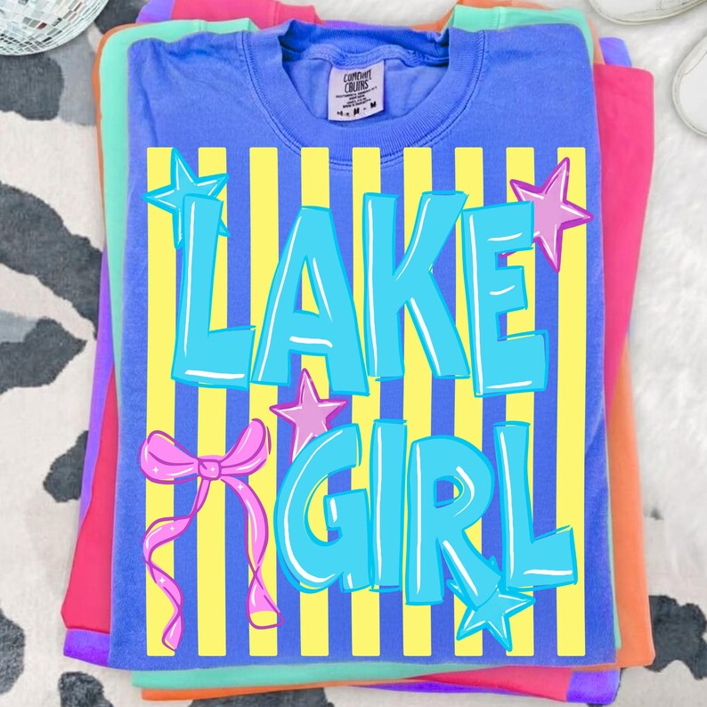 May include: A stack of colorful t-shirts. The top shirt is blue with the words "LAKE GIRL" in turquoise, set against yellow vertical stripes. Pink stars and a bow accent the design. The shirts underneath are pink, green, and orange.