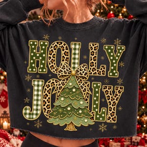 May include: A charcoal gray long-sleeve sweatshirt featuring the text HOLLY JOLLY in a mix of green polka dot, leopard print, and green gingham patterns. A central graphic shows a green Christmas tree topped with a large leopard print bow.