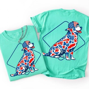 Puede incluir: Una camiseta verde menta con el gr&aacute;fico de un perro que lleva una gorra de b&eacute;isbol. El perro tiene un patr&oacute;n de estrellas, rayas y huellas en rojo, blanco y azul. Un contorno de diamante azul oscuro enmarca al perro y una cadena plateada.