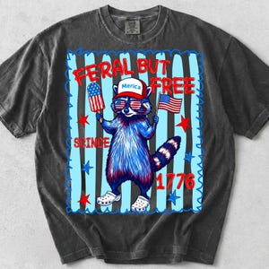 May include: A charcoal grey t-shirt featuring a graphic of a raccoon wearing a hat that says Merica, sunglasses with American flag lenses, and white clogs. The raccoon holds a popsicle and a flag. Text reads FERAL BUT FREE SINCE 1776.