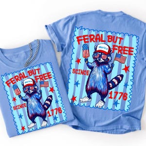 May include: A light blue t-shirt featuring a graphic of a raccoon wearing a red hat with the text Merica, sunglasses with American flag patterns, and white clogs. The raccoon holds a popsicle and a flag, with the text Feral But Free Since 1776.